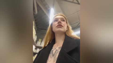 Media: Video of a blonde woman with a tattoo on her chest, wearing a black blazer, in a dimly lit, industrial-looking room with exposed pipes and a ceiling fan.