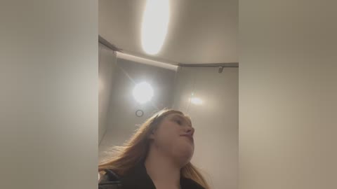 Media: Video of a woman with long, light brown hair, wearing a black coat, standing inside a dimly lit, cylindrical elevator. Her eyes are closed, and her head is tilted back slightly.