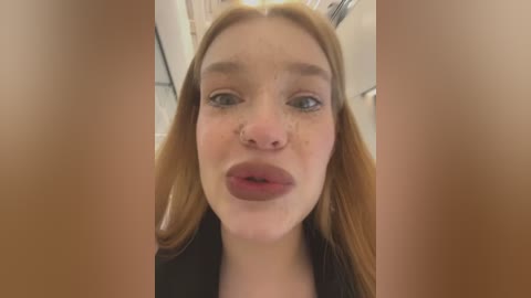 Media: Video of a young Caucasian woman with fair skin, freckles, long auburn hair, and a septum piercing. She has full lips and wears a black top.