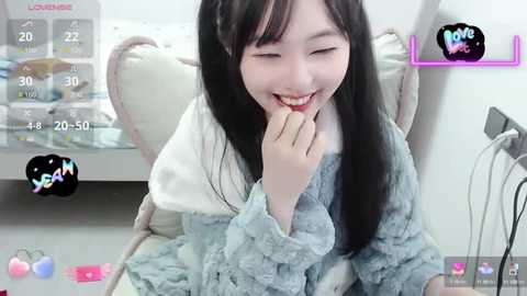 Media: A video of a young Asian woman with long black hair, smiling, with her finger to her lips, in a cozy room with white walls and a bed.