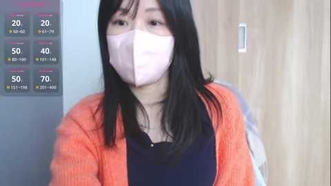 Media: A video of an Asian woman with long black hair, wearing a pink face mask and an orange cardigan over a navy top, set against a wooden backdrop.