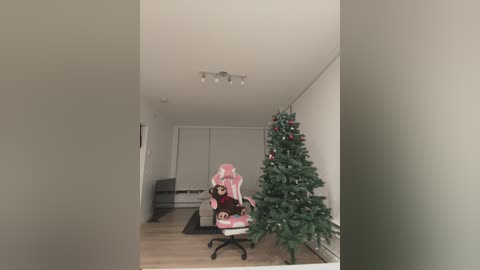 A video of a modern, minimalist living room with a pink gaming chair, a decorated Christmas tree, and a sleek gray couch, featuring a plush teddy bear.