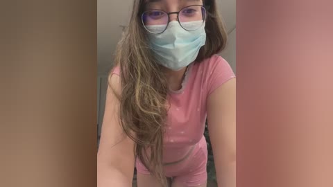 Media: Video of a young Asian woman with long brown hair, wearing a blue surgical mask, black-rimmed glasses, and a pink short-sleeve shirt. She leans forward, captured from above, in a sterile room with beige walls.