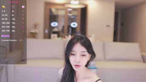 Media: Video of an Asian woman with long black hair, wearing a black top, sitting in a modern living room with a gray couch, wooden door, and a chandelier.