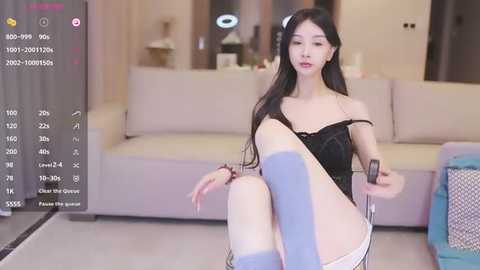 Media: Video of an East Asian woman with long black hair, fair skin, wearing a black lingerie top, knee-high blue socks, and sitting provocatively on a couch.