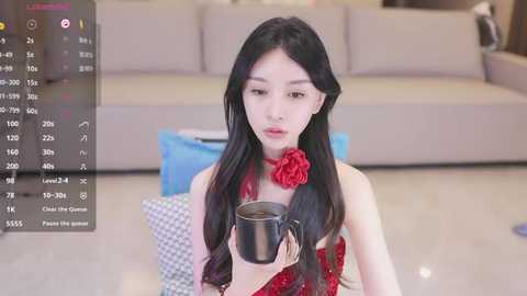 Media: Video of a young Asian woman with long black hair, wearing a red dress with a red rose, holding a coffee mug, indoors on a light-colored floor, with a modern grey sofa in the background.