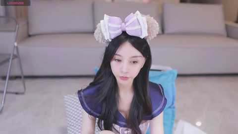 Media: Video of a young Asian woman with long black hair, wearing a sailor outfit and white bunny ears. She sits on a light-colored floor, in a modern living room with a gray sofa in the background.