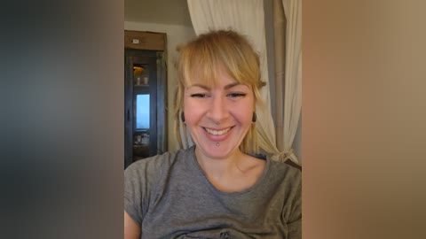 Media: Video of a smiling, light-skinned, blonde woman with bangs, wearing a grey T-shirt, standing in a dimly lit room with beige walls, a wooden door, and a window.