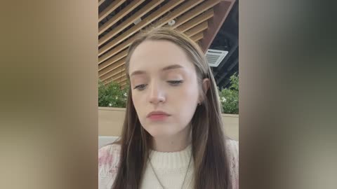 Media: A video of a young Caucasian woman with long, straight brown hair, wearing a white sweater, under a wooden slatted roof. The background features a green leafy plant and a ceiling fan.