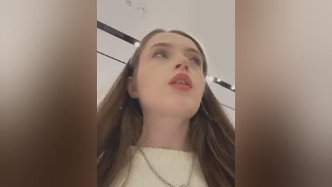 Media: Video of a young woman with long brown hair, wearing a cream sweater, captured from a low angle. Her face is slightly tilted upward, showing a neutral expression. The background is a modern, minimalist room with white walls and ceiling lights.