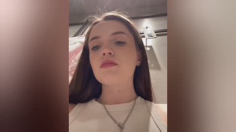 Media: Video of a young woman with fair skin and long brown hair, wearing a white top and a silver necklace, indoors with blurred background of a bedroom.