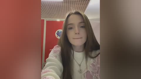 Media: A video of a young, fair-skinned woman with long brown hair, wearing a white sweater and a pink lace cardigan, smiling in a brightly lit room with red and white walls and a grid ceiling.