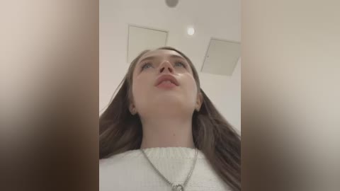 Media: Video of a young Caucasian woman with long brown hair, fair skin, and a neutral expression, wearing a cream-colored sweater and a silver necklace. The background features a modern, minimalist interior with white walls and recessed lights.