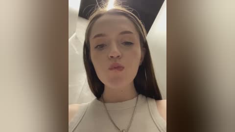 Media: Video of a young Caucasian woman with long brown hair, wearing a white tank top, standing indoors with a blurred background, making a kissy face.