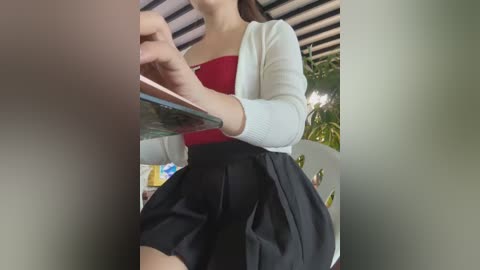 Media: A video of a young Asian woman with fair skin, wearing a red strapless top and a white cardigan, sitting outdoors with a black pleated skirt.