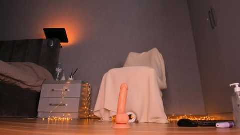 Media: Video of a dimly-lit bedroom with a grey nightstand, a white chair, a dildo on the floor, fairy lights, and a warm orange lamp casting a cozy glow.