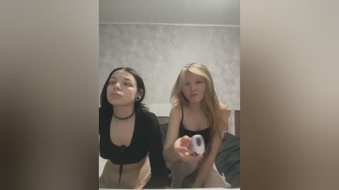 Media: A video captures two young women in a dimly lit room. One with black hair, wearing a black top, and another with blonde hair, in a black top, both smiling and holding a toy.