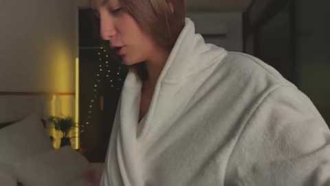 Media: A video of a woman with light skin, brown hair, and a white towel, coughing, standing indoors near a lamp and a bed.