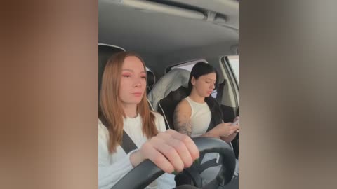 Media: Video of two young women in a car, one with light skin and straight brown hair, the other with medium skin tone and dark hair, both wearing white tops, looking at phones.