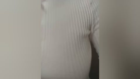 Media: A video of a person wearing a white, ribbed, long-sleeved t-shirt, standing against a plain, beige wall. The image is slightly blurry, focusing on the upper body and torso.