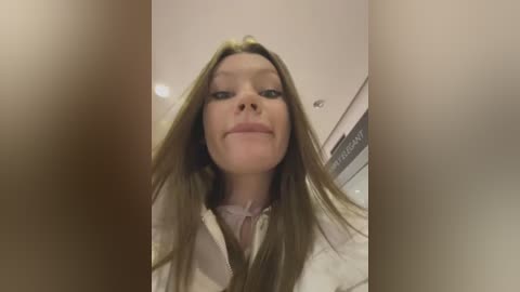 Media: A video of a smiling woman with long, straight brown hair, wearing a white hoodie, taken from a low angle, creating a distorted, wide-eyed effect. The background features a blurred, beige wall with a ceiling light and a partially visible sign.