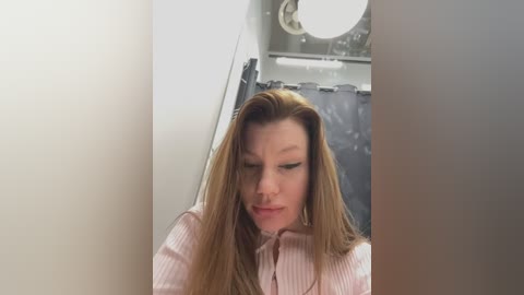 Media: A video of a young Caucasian woman with long, straight blonde hair, wearing a pink striped shirt, standing in a bathroom with a shower curtain and white walls.