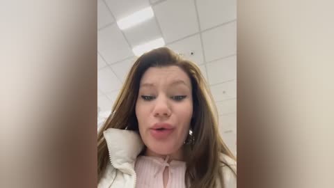 Media: Video of a woman with long brown hair, wearing a light pink blouse, puckering her lips in a selfie taken from below, in a brightly lit office with white ceiling tiles and fluorescent lights.