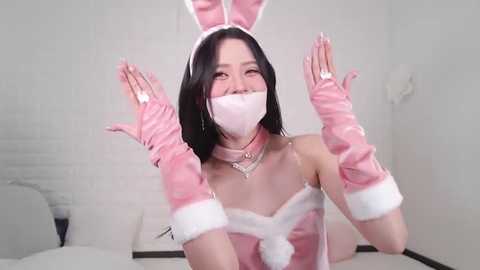 Media: Video of an Asian woman with long black hair, wearing a pink bunny-themed outfit, including a headband, gloves, and a mask, smiling with hands up.