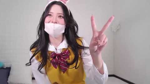 Media: Video of a young Asian woman with long black hair, wearing a yellow vest over a white shirt, plaid bow tie, and white mask, making a peace sign.