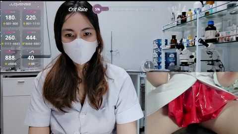 Media: Video of an Asian woman in a white lab coat and mask, standing in a sterile lab with a glass shelf filled with medical supplies and bottles, and a chart showing COVID-19 statistics.