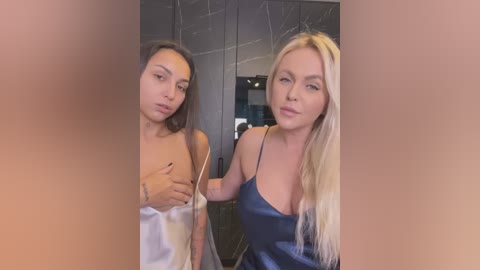 Media: Video of two women in a bathroom: a slender, dark-haired woman with a white towel covering her chest, and a blonde woman in a blue satin camisole, both smiling.