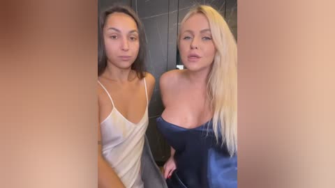 Media: Video of two women, one with long brown hair in a white top, the other with long blonde hair in a dark blue top, standing side-by-side in a dimly lit bathroom.