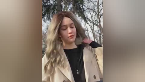 Media: Video of a young woman with long, wavy, light blonde hair, wearing a beige trench coat over a black top, standing outdoors in a park with bare trees and cloudy sky.