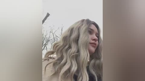 A video of a young woman with long, wavy, platinum blonde hair, wearing a beige coat, standing against a foggy background with bare tree branches visible.