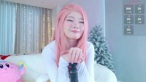 Media: Video of a young Asian woman with long pink hair, wearing a white sweater, sitting on a couch with a plush toy beside her, smiling.