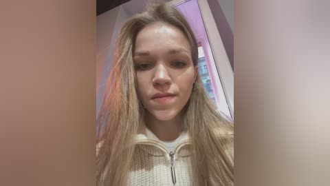 Media: Video of a young Caucasian girl with long, light brown hair, wearing a beige zip-up hoodie, looking slightly sad in a dimly lit room with blurred, pastel-colored walls.