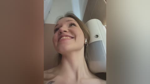 Media: Video of a topless woman with fair skin and blonde hair, smiling and looking upwards, inside a white-tiled bathroom with a white wall-mounted air purifier.