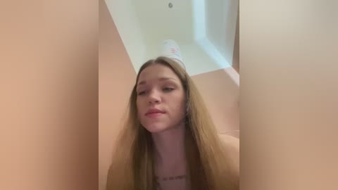 Media: Video of a young Caucasian woman with long, straight blonde hair, wearing a beige top, captured from a low angle, looking slightly downcast in a small, dimly lit room with beige walls and a white ceiling.