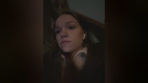 Media: Video of a young woman with light skin and long brown hair, wearing a brown coat, lying on a dark couch, looking into the camera with a neutral expression.