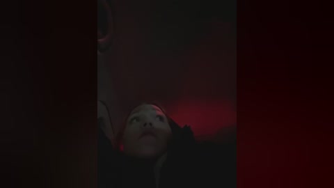 Media: A dimly lit, atmospheric video shows a woman with pale skin and dark hair, lying on her back, her face illuminated by a red light, creating a dramatic, eerie mood.