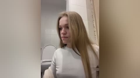 Media: A video of a young Caucasian woman with long, straight blonde hair, wearing a light blue sweater, standing in a small, white-tiled bathroom with a closed toilet and beige walls.