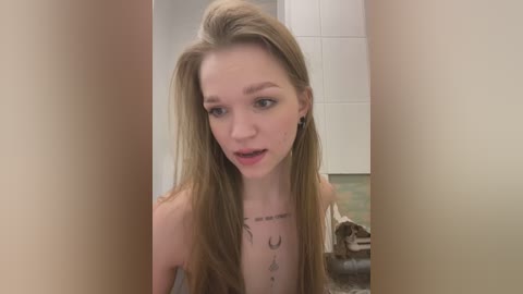 Media: Video of a topless, fair-skinned woman with long blonde hair, wearing black earrings, and a necklace with text, standing in a bathroom with white tiles and a green shower curtain.