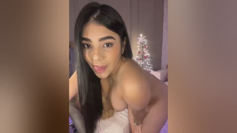 Media: A video of a young woman with long black hair, light skin, and a slender physique, smiling, partially topless, in a dimly lit bedroom with a Christmas tree.