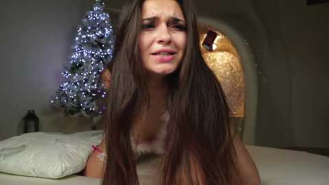 Media: Video of a young woman with long brown hair, wearing a Santa hat, crying on a bed with a Christmas tree in the background, dimly lit room.