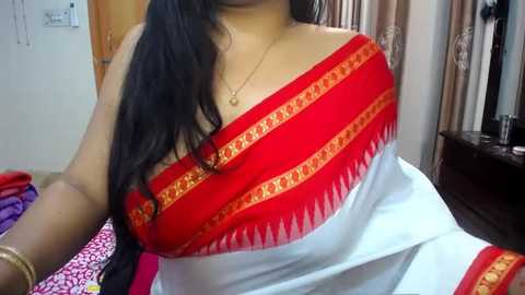 Media: A video of a woman wearing a red and white saree with gold embroidery, seated indoors, showing her upper body. She has long black hair, wearing gold bangles and a necklace.