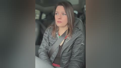 Media: Video of a young woman with straight, shoulder-length brown hair, wearing a dark grey puffy jacket and a black top, sitting in a car with a seatbelt. The background shows a blurred interior with a steering wheel.