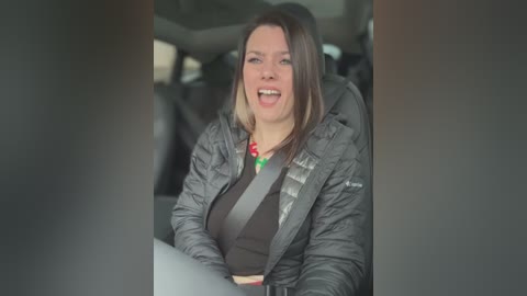 Video of a middle-aged woman with straight brown hair, wearing a gray puffer jacket and a red necklace, seated in a car with a safety belt on. She is smiling broadly, her face animated.