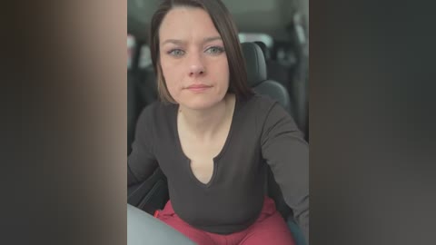 Media: Video of a Caucasian woman with short brown hair, wearing a black V-neck top and red pants, sitting in a car.