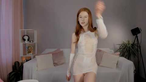 Media: A video of a slender, red-haired woman in a sheer, white lace dress waving in a modern living room with grey walls, white couch, and potted plants.