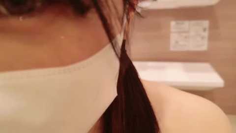 Media: Video of a woman's bare back, with long dark brown hair pulled to one side, in a beige tiled bathroom with a white toilet and sink visible in the background.